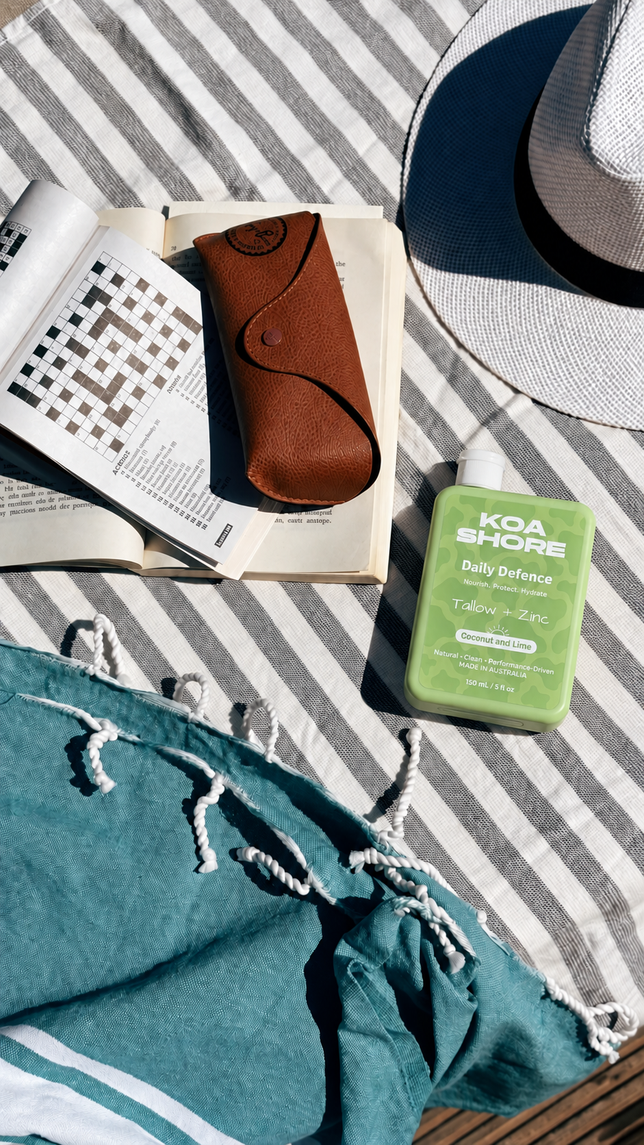 Coconut and Lime Daily Defence beside a book and sunglasses on a striped beach towel